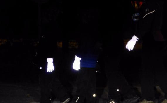 Heijastavat toppahanskat koko perheelle. Pimeässä aikuinen ja kaksi lasta ja kaikilla heijastavat toppahanskat kädessä.