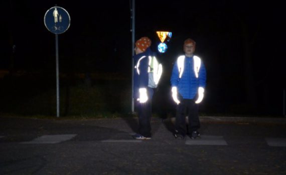 Kaksi lasta pimeällä suojatiellä. Selässä heijastavat reput ja käsissä heijastavat toppahanskat. Hyvin näkyvät pimeällä.